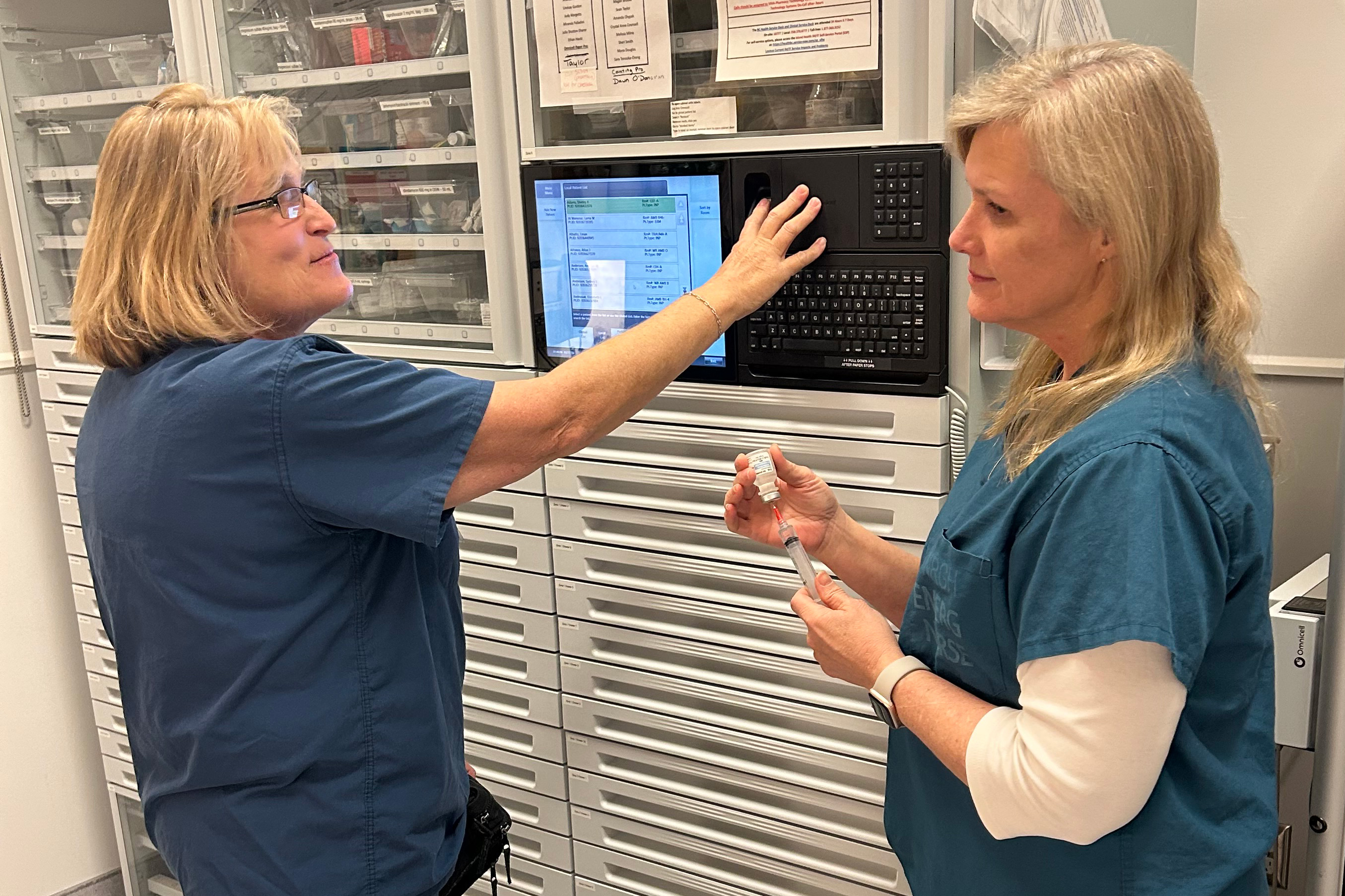 Two nurses converse with one another in front of a medication dispensing machine at a hospital.