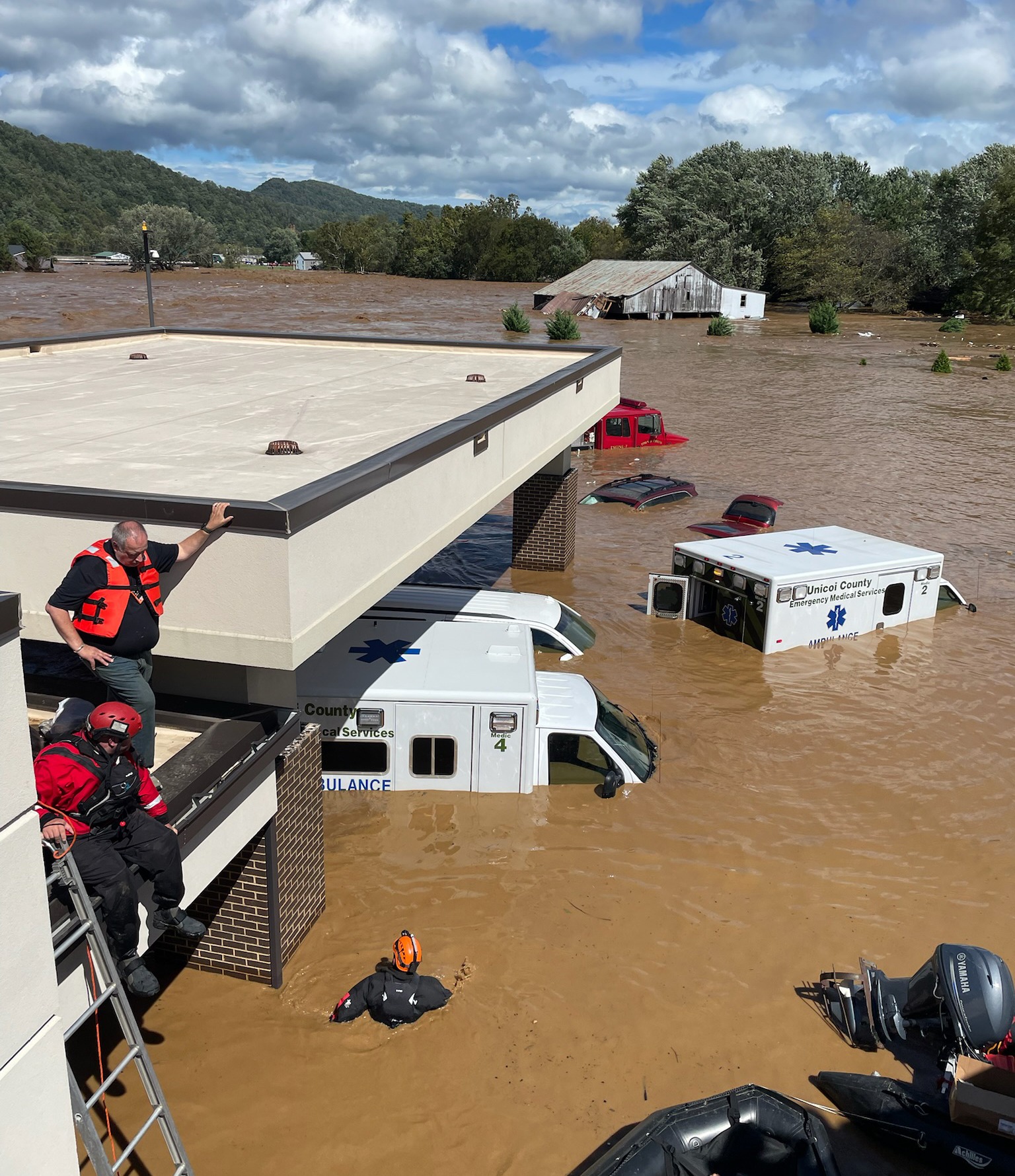 Unicoi County Hospital during the flood. Ambulances are nearly submerged in muddy water. Two people in red vests are seen climbing on to the roof.