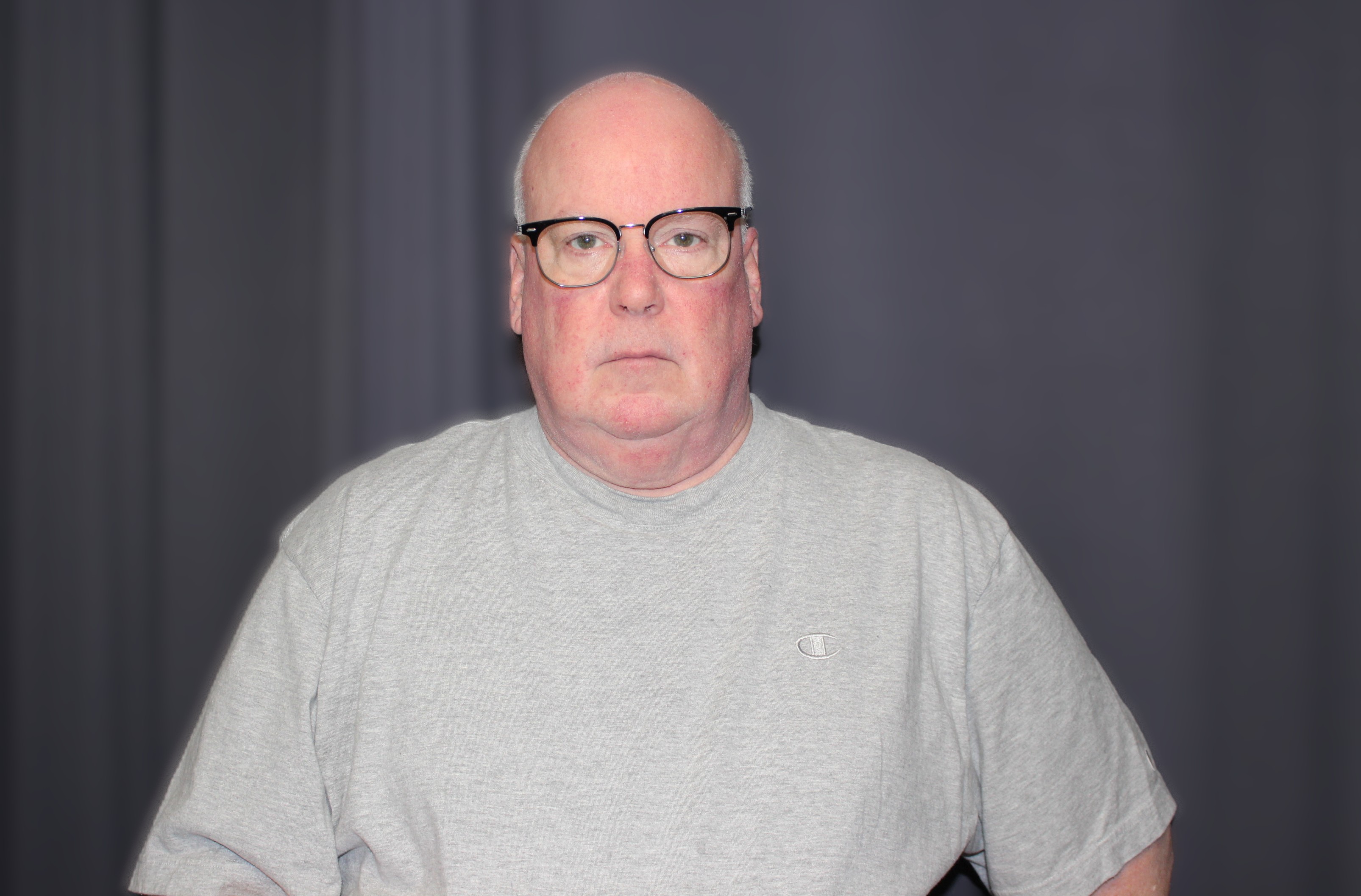 An older man with glasses stands for a photo against a dark gray backdrop.