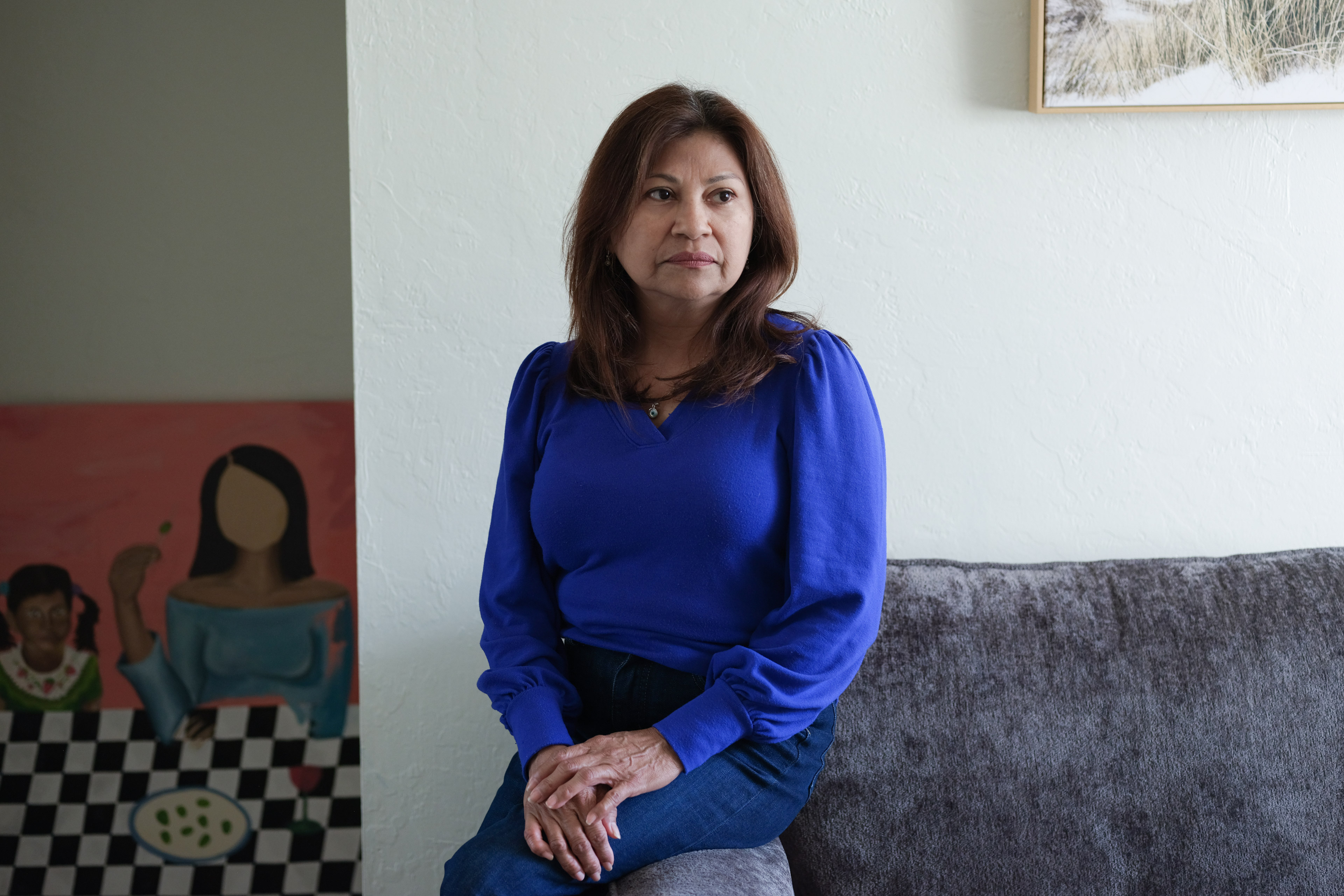 Lorena Alvarado Hill sits on the edge of her couch. A mural painting is seen on the wall behind her.