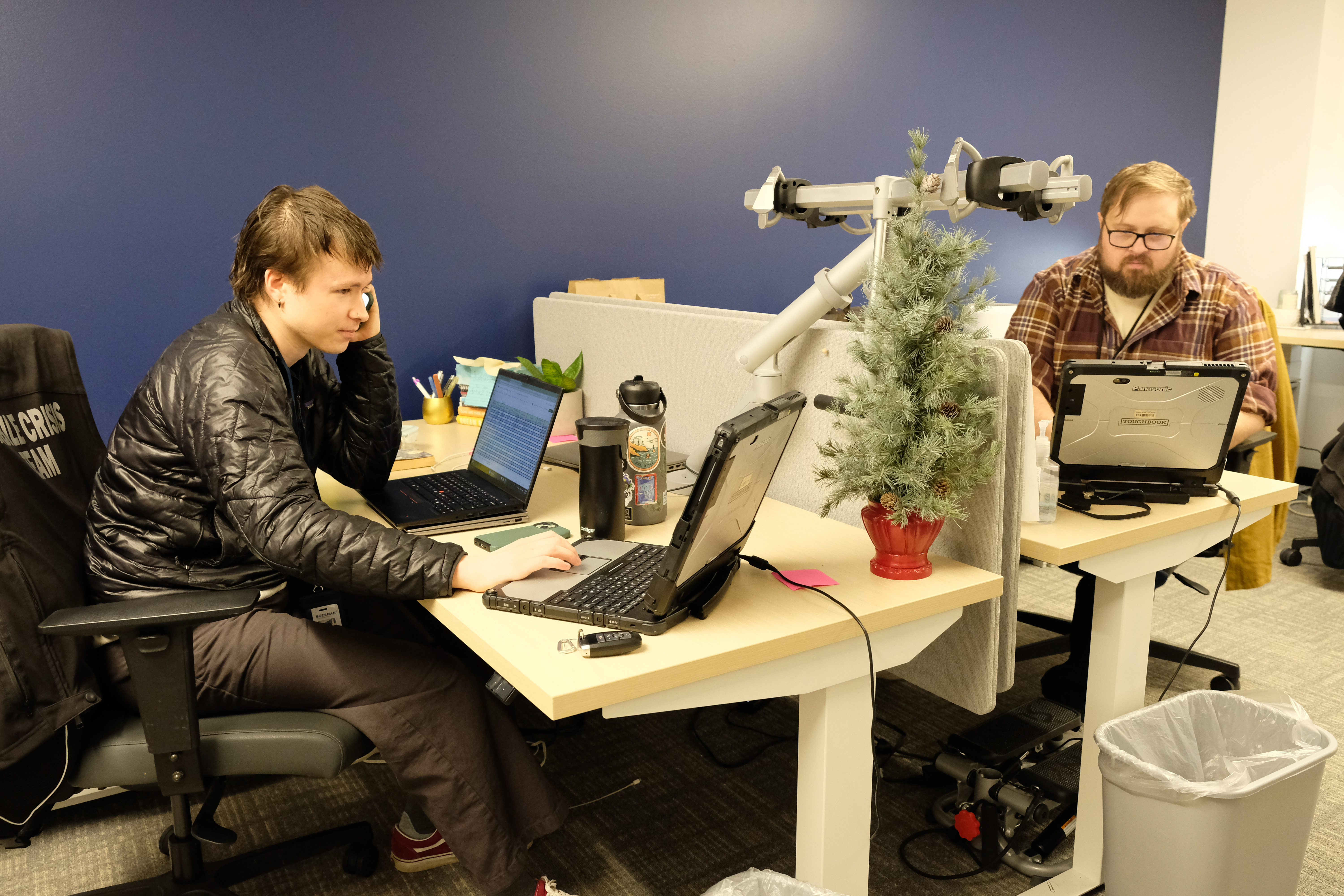 Two men are seated at a white, divided desk with a small christmas tree on the table 