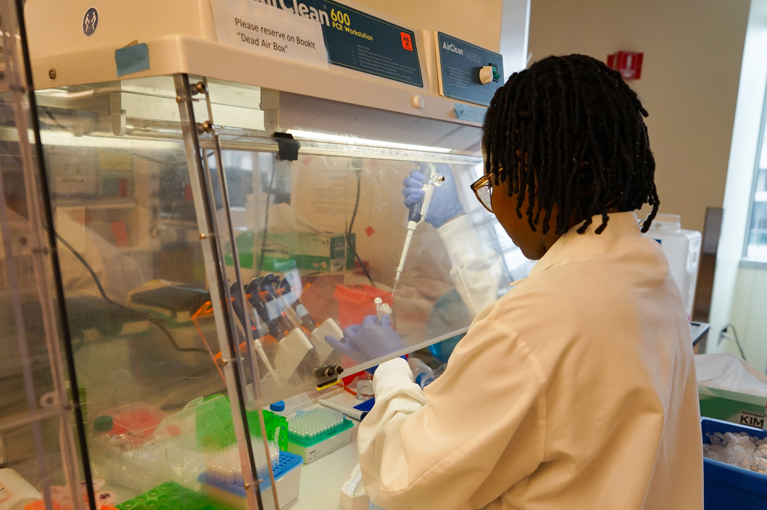 A Black woman in a labcoat works with a laboratory pipette, her hands shielded behind a pane of glass.