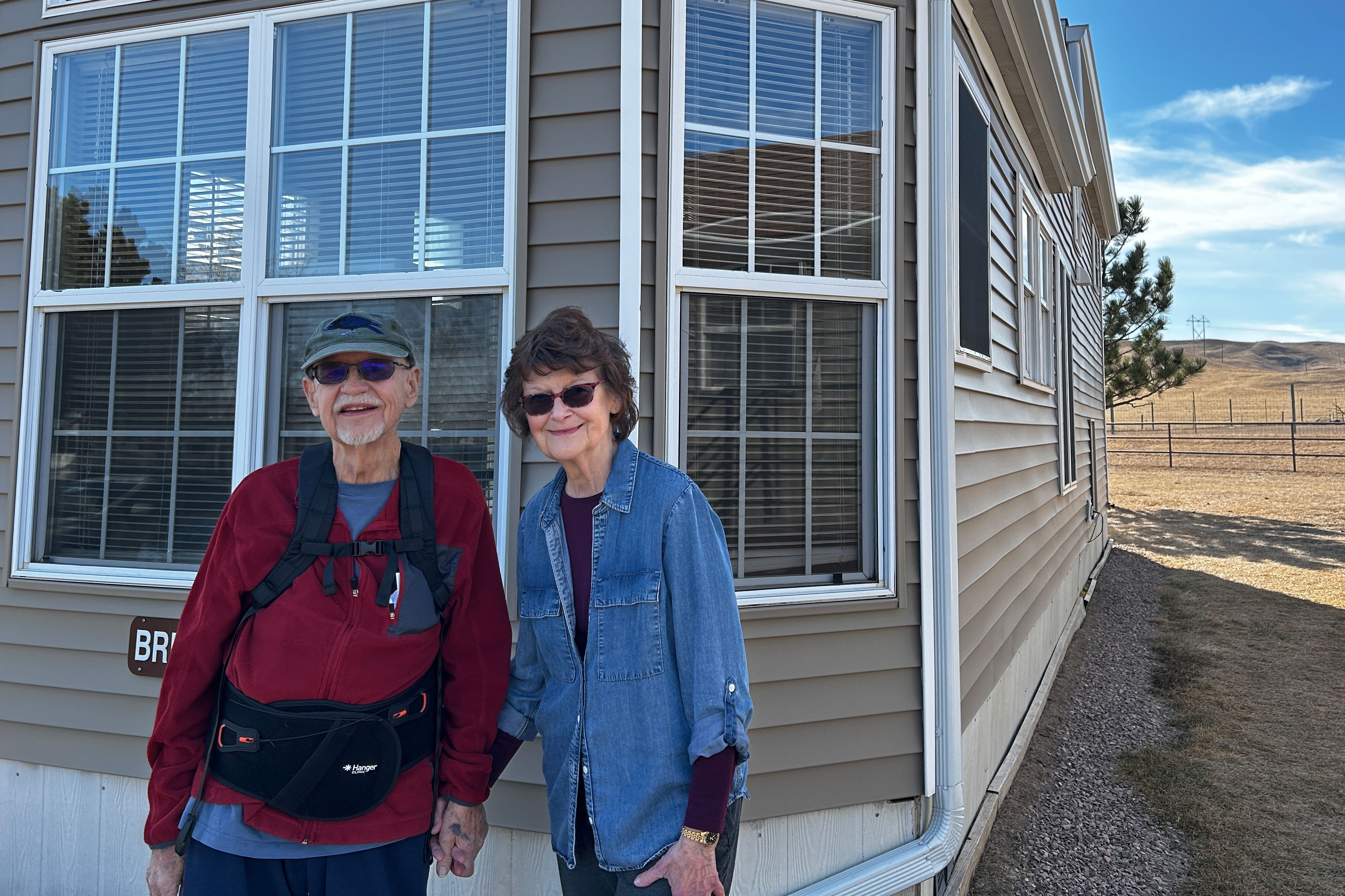 An older couple stand outside a beige-colored house.